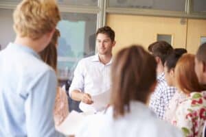 teacher-with-college-students-standing-by-desks-PZL39MX