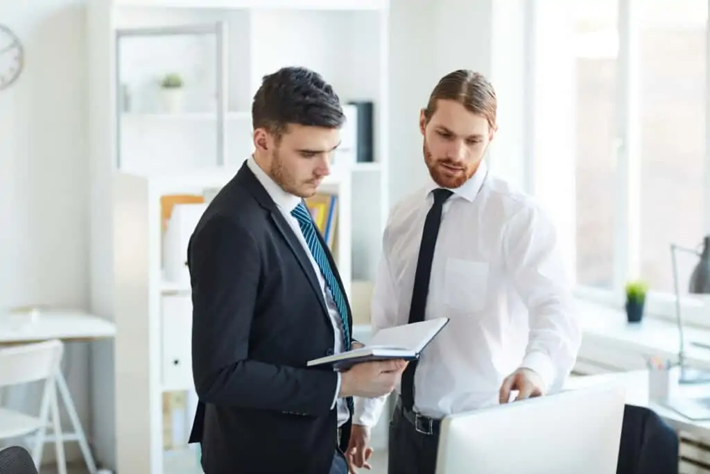 Two young brokers in formalwear standing by computer monitor in office and discussing online information