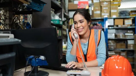 smiling asian young female using telephone talking with client at warehouse factory office. woman worker using desktop computer check product order shipment for customer.